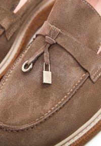 Gros plan sur un mocassin en daim marron avec un gland décoratif en cuir et un petit cadenas argenté sur le dessus.