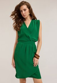 Grünes Wickelkleid mit tiefem V-Ausschnitt, ärmellosem Design, geraffter Taille und einem fließenden Rock. Das Model trägt Holzarmreifen und hat wellige Haare.