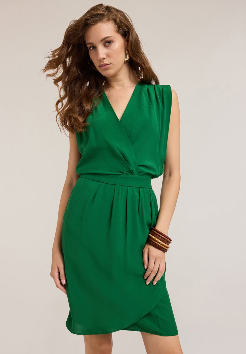 Grünes Wickelkleid mit tiefem V-Ausschnitt, ärmellosem Design, geraffter Taille und einem fließenden Rock. Das Model trägt Holzarmreifen und hat wellige Haare.