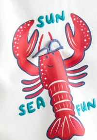 Rød tegneseriehummer med broderede blå og hvide ører på hvidt stof, omgivet af turkis broderede ord "SOL," "HAV" og "SJOV."