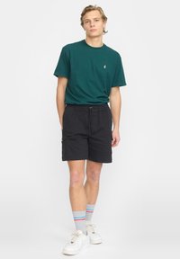 Jeune homme debout portant un t-shirt vert foncé, un short cargo noir, des chaussettes grises avec des rayures rouges et bleues, et des baskets blanches sur fond blanc.