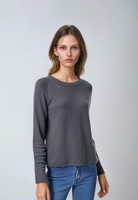 Szary sweter o dzianinowej strukturze z okrągłym dekoltem, prążkowanymi krawędziami i długimi rękawami, połączony z niebieskimi dżinsami. Gładka textura z delikatnymi poziomymi liniami.