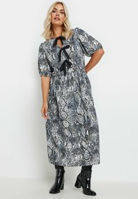 Pitkähihainen, puffihihallinen mekko harmaassa ja mustassa käärmeennahkakuosissa, jossa on solmittava kaulakoriste ja vartalonmyötäinen vyötärö, hulmuava hame sekä mustat nilkkurit.