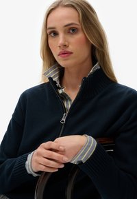 Pull-over marine avec col montant et fermeture éclair, superposé sur une chemise à rayures bleu et blanc. Texture tricotée avec des poignets côtelés.