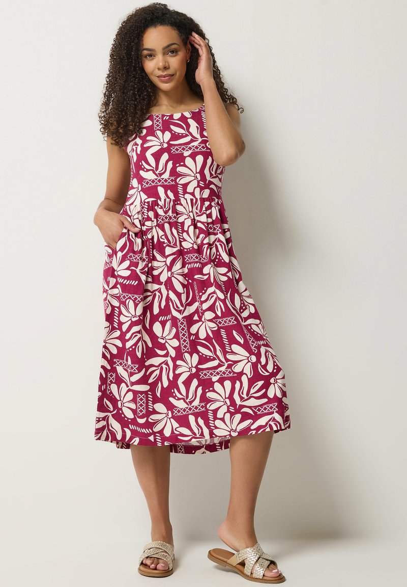 Mujer con cabello rizado, vistiendo un vestido sin mangas hasta la rodilla de color granate con grandes patrones florales blancos y sandalias trenzadas beige.