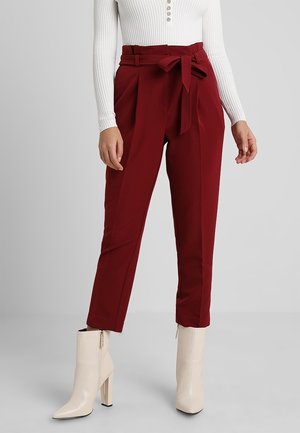 Woman wearing high-waisted, wine-red paperbag pants with a tied belt, paired with white heeled ankle boots and a white ribbed long-sleeve top.