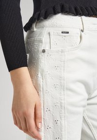 Beli vezeni jeans iz denima s cvetličnim vzorci, sprednjim gumbom in prilegajočim se dizajnom, v kombinaciji s črno rebrasto majico.