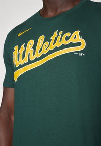 Zelené bavlněné tričko s žlutým nápisem „Athletics“ s bílým obrysem. Ikonické logo Nike v žluté barvě na rameni. Měkká textura.