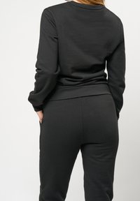 En person i svart sweatshirt och byxor står med ryggen vänd mot kameran, med händerna i fickorna, mot en ljus bakgrund.