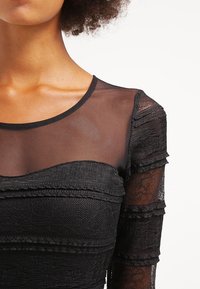 Robe noire avec des manches en maille transparente et un corsage texturé avec du tissu superposé. Le design comprend des accents plissés et un col rond.