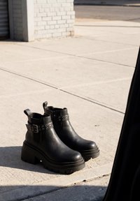 Ζευγάρι μαύρα δερμάτινα chunky μπότες με πλατφόρμα, λουρί και λεπτομέρεια πόρπης πάνω σε τσιμεντένιο πεζοδρόμιο στο φως της ημέρας.