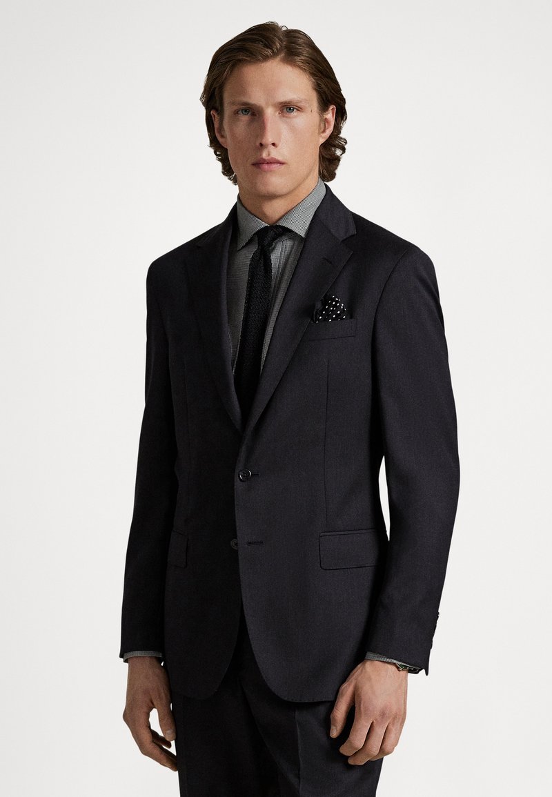 Ciemny, dopasowany garnitur z teksturowaną, szarą koszulą i czarnym krawatem. Charakterystyczna poszetka w kropki. Gładka tkanina o usztywnionym kroju.