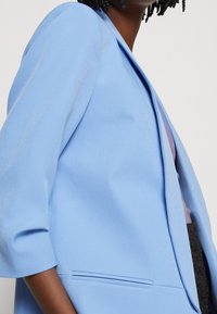 Woman wearing a light blue blazer with three-quarter sleeves, showing part of a lavender top and dark textured pants.