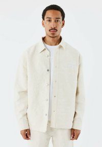 Beige mönstrad skjortjacka med knäppning framtill, med synliga sömmar, en subtil struktur och en liten broderad logotyp på bröstet.