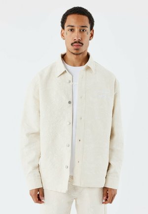 Beige gebloemde overhemdjas met een knoopsluiting aan de voorkant, voorzien van zichtbare stiksels, een subtiele textuur en een klein geborduurd logo op de borst.
