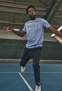 Camisa deportiva azul claro con el logo "BOSS", pantalones negros y zapatillas blancas en una cancha de tenis, mostrando un movimiento dinámico.