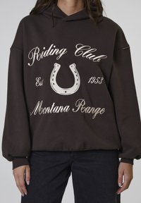 Persoon in een donkerbruine hoodie met de tekst "Riding Club Montana Range Est 1953" en een hoefijzer graphic op de voorkant.