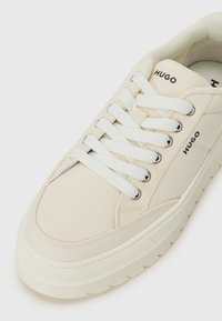 Beige canvas sneaker with white laces, featuring silver eyelets and a textured sole. Branding "HUGO" is visible on the sides.