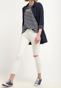 Marineblaue Langarmjacke über einem marineblauen und weißen gestreiften T-Shirt. Hellblaue Skinny-Jeans mit Rissen an den Knien, kombiniert mit marineblauen Sneakers.
