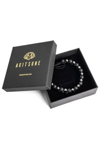 Black gift box with gold logo, containing a beaded bracelet with black lava stones and shiny metallic accents, inside a black interior.