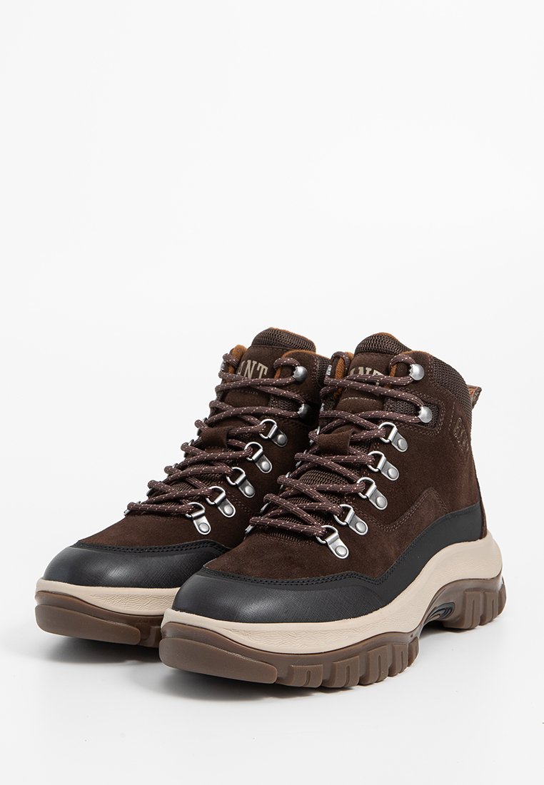 Een paar bruine hiking schoenen met metalen veterogen, dikke beige zolen en zwarte neuzen op een witte achtergrond.