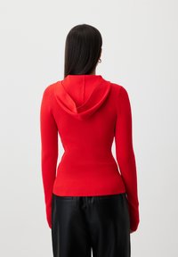 Red ribbed hoodie with a fitted design and long sleeves. Features a soft texture and a prominent drawstring hood. Worn with black trousers.