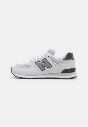 Jasnoszary sneaker New Balance z czarnymi akcentami, białymi sznurowadłami i amortyzowaną podeszwą na białym tle, widok z boku.