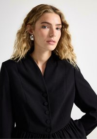 Kvinde med bølget blond hår iført en sort knappet blazer og store sølvøreringe, stående mod en ensfarvet hvid baggrund.