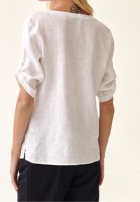 Donna che indossa una camicia bianca di lino larga con le maniche arrotolate e pantaloni neri, mostrando un tatuaggio sul collo su uno sfondo chiaro e semplice.