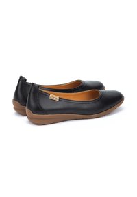 Ballerines noires en cuir avec un bout rond, semelle en caoutchouc beige et une étiquette de marque discrète. Texture lisse et design simple.