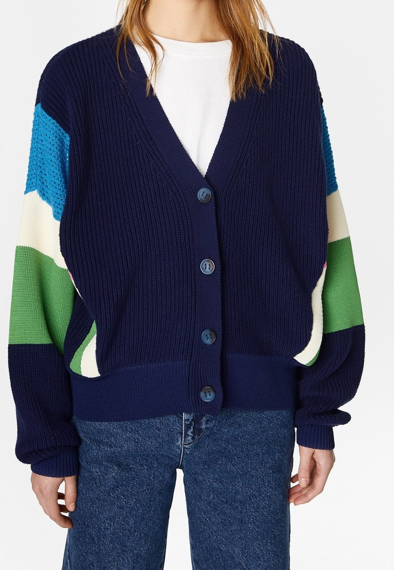 Cardigan en maille bleu marine avec une coupe loose, présentant des manches à motifs colorés en bleu, vert et crème, fermeture par boutons, poignets côtelés.