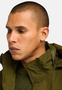 Chaqueta verde oliva con cuello alto, cierre con cremallera y costuras visibles. Capa sobre un suéter de punto verde texturizado. Primer plano de la parte superior del cuerpo.