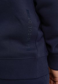 Granatowy sweter z teksturowanym ścięciem u dołu i po bokach, z wytłoczonym napisem "BLUE RIDGE DENIM" wzdłuż lewej strony. Miękka powierzchnia materiału.