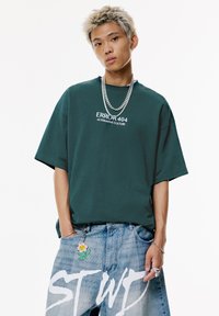Green oversized T-shirt with "ERROR 404 ALTERNATIVE CULTURE" printed in white; paired with light blue distressed jeans featuring a flower keychain.
