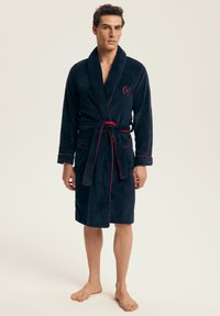 Mężczyzna stojący boso, ubrany w granatowy szlafrok z czerwonymi wykończeniami, z dwiema przednimi kieszeniami i przewiązaną pętelką na jednolitym jasnym tle.