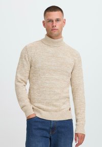 Joven con cabello corto que lleva un suéter de cuello alto beige y jeans azules, posando con una mano en el bolsillo frente a un fondo blanco.