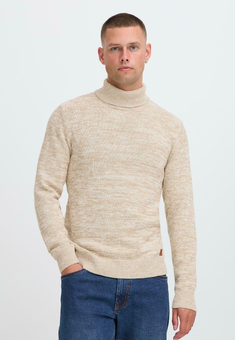 Joven con cabello corto que lleva un suéter de cuello alto beige y jeans azules, posando con una mano en el bolsillo frente a un fondo blanco.