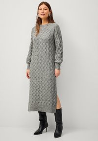 Robe en maille grise avec un motif en relief, manches longues et fente sur le côté, assortie à des bottes à talons noires.