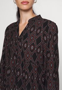 Svart blus med mönster som har en diamantdesign i röd, vit och blå. Tyget har en strukturerad yta och en V-ringning.