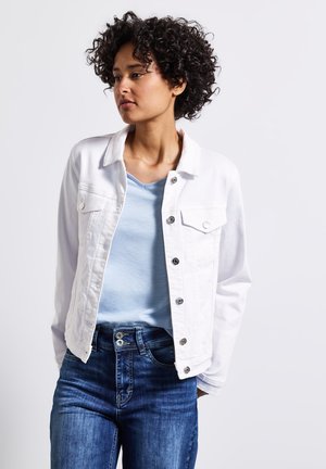 Veste en denim blanc avec fermeture à boutons et deux poches poitrine, associée à un haut bleu clair à col en V et un jean foncé ajusté. Cheveux bouclés.