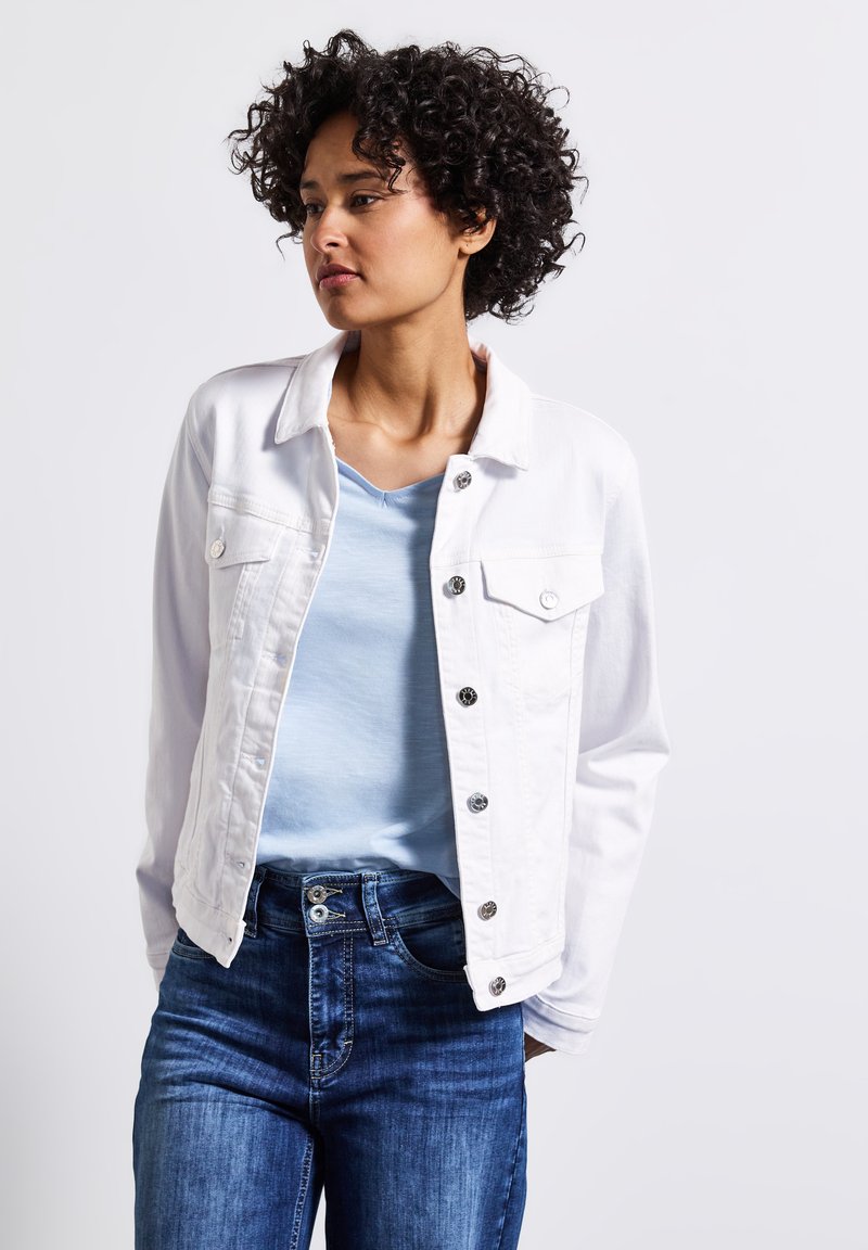 Biała dżinsowa kurtka z zapięciem na guziki i dwoma kieszeniami na piersi, w połączeniu z jasnoniebieskim topem w serek i ciemnoniebieskimi dopasowanymi dżinsami. Kręcone włosy.