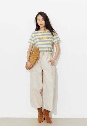 Jeune femme aux longs cheveux foncés porte un t-shirt rayé jaune et gris « amor », un pantalon large beige, des bottes en suède marron et tient un sac marron assorti.