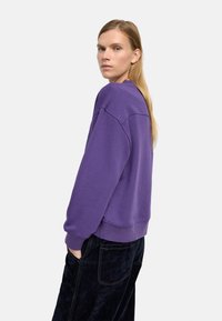 Fioletowa bluza z luźnym krojem, z prążkowanymi mankietami i dołem, noszona na ciemnych, fakturowanych spodniach. Prosty design bez widocznych wzorów czy logo.