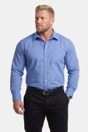 Mand med kort blondt hår og skæg iført en blå langærmet skjorte med knapper, sort bælte, sorte bukser og et armbåndsur.