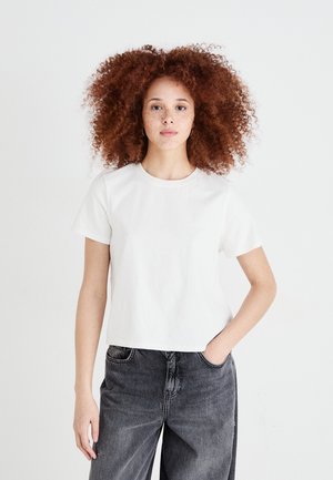 Witte katoenen T-shirt met korte mouwen en een ronde hals, gecombineerd met hoogwijn donkere grijze jeans. Het model heeft krullend rood haar en staat.