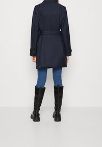 Trench coat azul marino con un cinturón, de longitud media, diseño entallado, combinado con jeans azules y botas negras hasta la rodilla. Tela suave, ajuste estructurado.