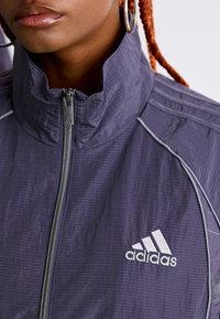 Személy lila Adidas cipzáras kabátot visel, a mellkasán a logó látható, nagy karika fülbevalóval és látható narancssárga fonott hajjal.