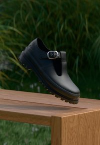 Black leather shoes with a T-strap design, a silver buckle, and rugged soles resting on a wooden surface against a green backdrop.