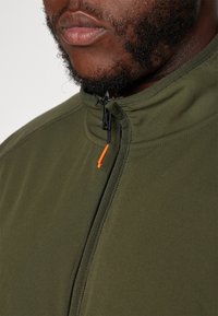 Primo piano del volto inferiore di un uomo con barba curata che indossa una giacca verde oliva con zip e un cursore della zip di colore arancione.