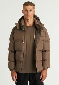 Chaqueta de abrigo marrón con capucha, que presenta puños elásticos, un cierre con cremallera y un sutil logotipo en el pecho. Lleva puesta una camiseta marrón.